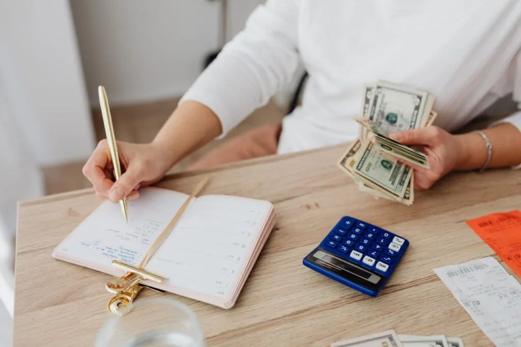 Adult holding cash and writing in planner while using a calculator for budgeting basics at home.