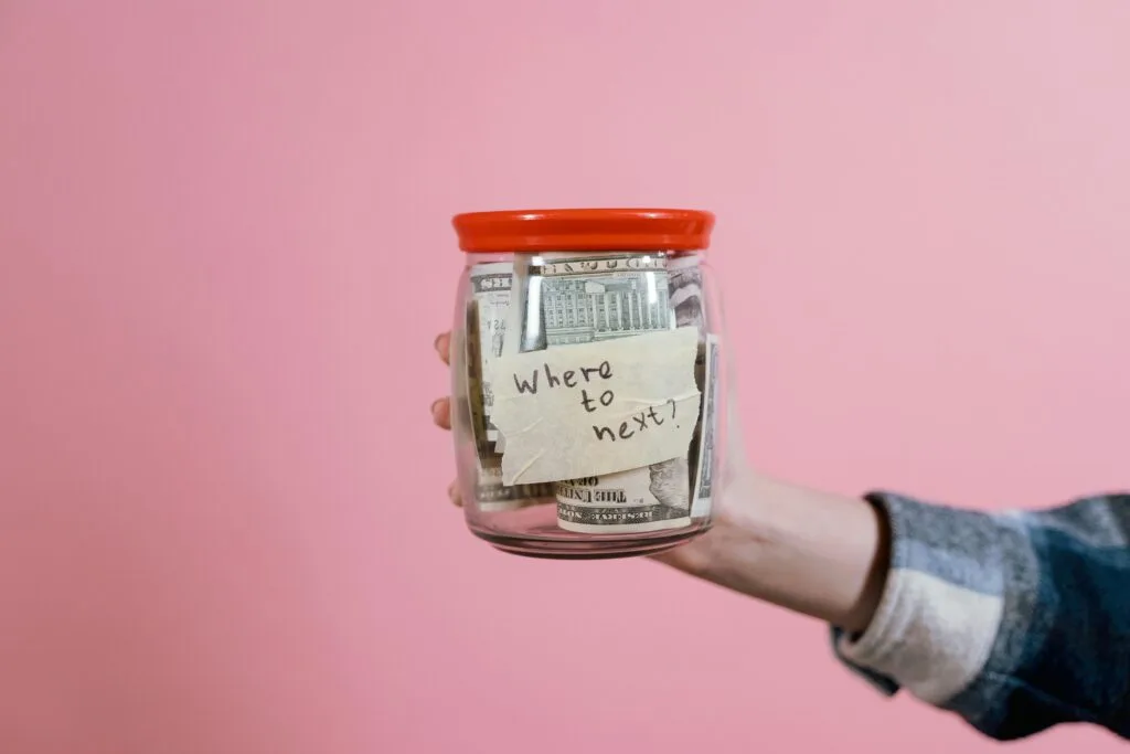 Vacation jar A hand holding a glass jar filled with dollar bills labeled 'Where to next?' against a pink background, symbolizing savings, and future planning.